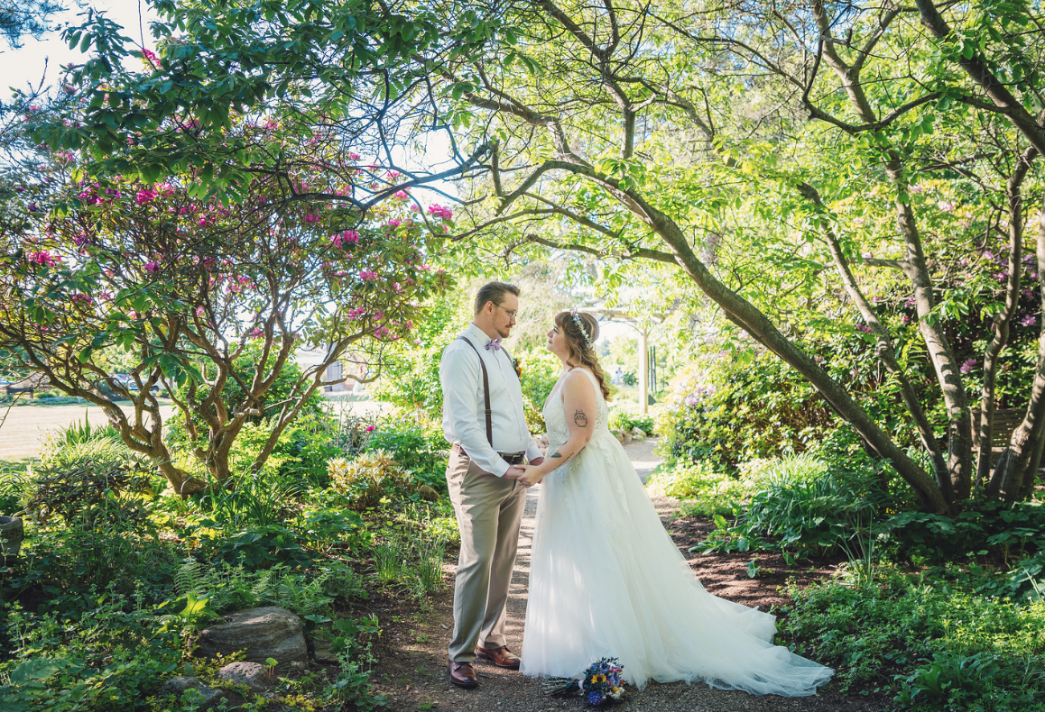 A Groom and Bride Hold Hands While Standing in a Beautiful Garden Pathway under trees